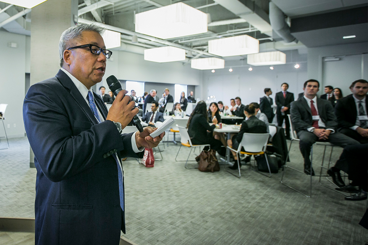 A man in a suit speaks into a microphone in front of a room full of people in business attire.