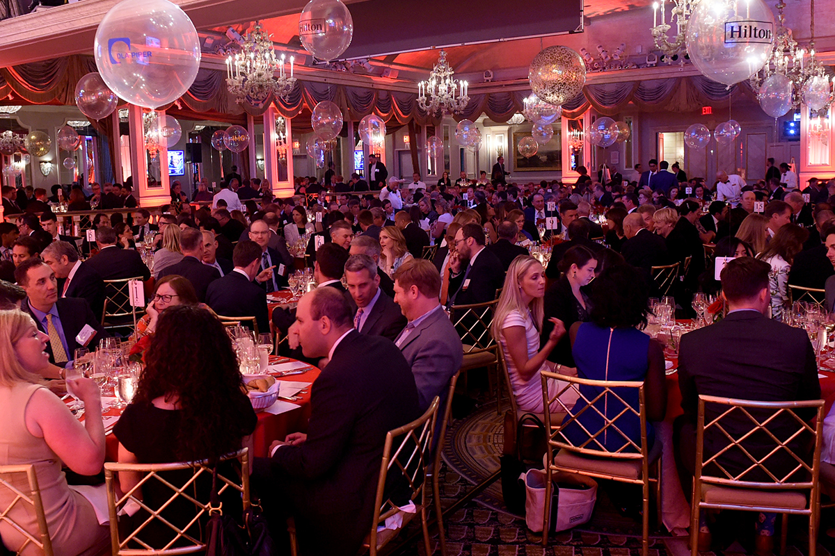 People in formal attire sit at round tables in a banquet room.