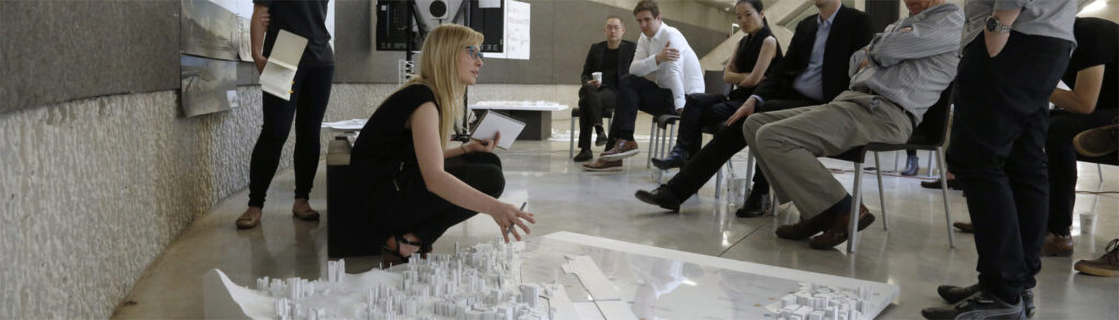 Student being down talking to forum of people in chairs presenting a white city model