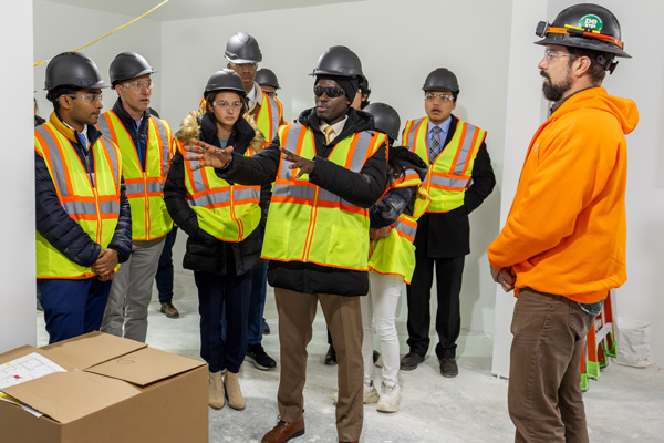 Students, wearing hard hats and construction vests, gather in an unfinished apartment; the student in the center is speaking.