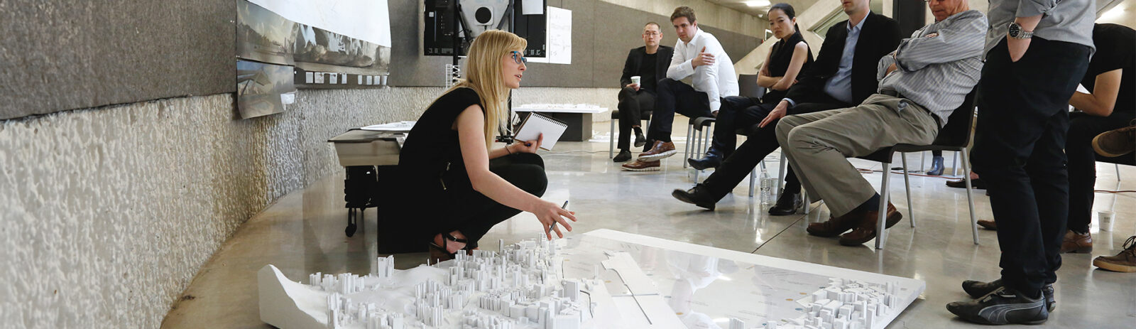 Professor bent down talking to forum of people in chairs presenting a white city model on the floor