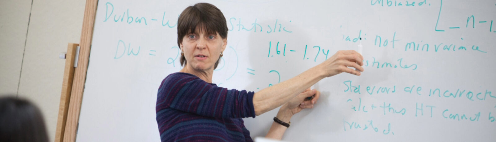 A woman stands in front of a whiteboard gesturing to the writing on the board.