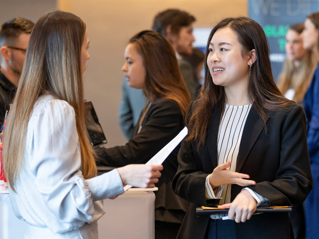 Two women in business attire speak to each other.